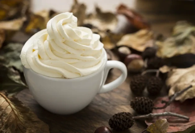 La imagen muestra una taza de café blanca repleta de crema batida, colocada sobre una superficie de madera rodeada de hojas secas otoñales, bellotas y pequeñas piñas, creando una atmósfera acogedora y estacional. La crema está esculpida en picos suaves, lo que sugiere una bebida preparada con cuidado y lista para ser disfrutada.