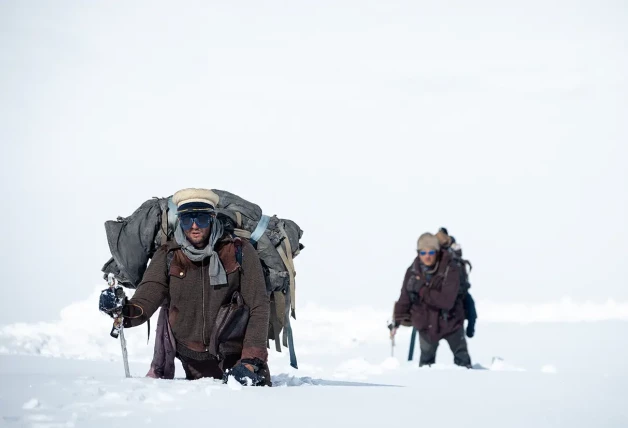 Escena de la película 'La sociedad de la nieve' con dos hombres andando por la montaña con la nieve hasta la cintura.