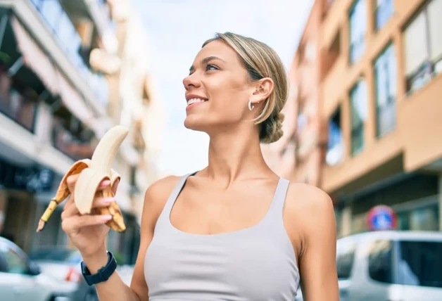 Joven mujer fitness usando ropa deportiva entrenamiento al aire libre comiendo plátano saludable para la fuerza y la energía