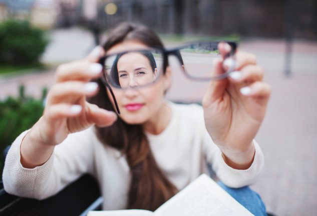 Closeup portrait of young women with glasses. She has eyesight problems and is squinting his eyes a little bit. Beautiful girl is holding eyeglasses right in front of camera with two hand.