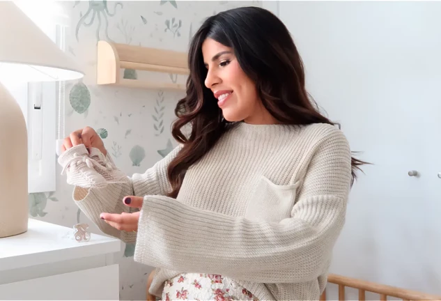 Isa Pantoja sonriente en la habitación de su futuro hijo con unos patucos en la mano