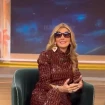 La imagen muestra a Marisa Martín-Blázquez sonriente sentada en un sillón verde, vestida con un traje estampado de color marrón y gafas de sol grandes. Ella sostiene un teléfono móvil en sus manos y parece estar en un entorno de plató de televisión, con un fondo que simula un cielo estrellado.