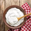 La imagen muestra un cuenco de barro lleno de nata montada (crema batida) sobre una mesa de madera rústica, acompañado de una servilleta a cuadros rojos y blancos y una cuchara de madera que descansa en la nata. La composición evoca un ambiente hogareño y tradicional.