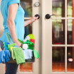 A house spring cleaning service person, a woman maid or cleaner arriving at the door entrance of customer home in the USA. She carries a cleaning kit of tools including rags, cleaning liquids, rubber gloves, and sponges. She wears casual clothes of t-shirt and jeans, arriving for clean-up work.