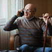 Anciano con bastón sentado en una butaca mirando por la ventana y hablando por teléfono.