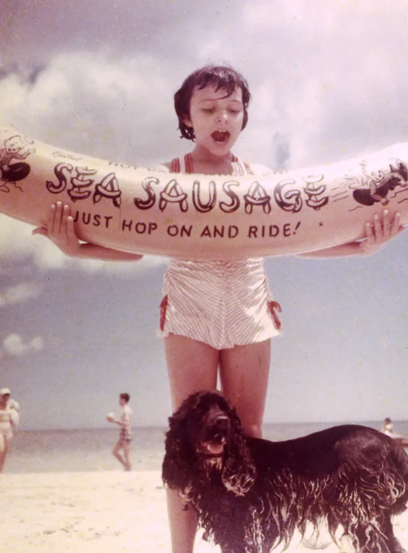 Mayra Gómez Kemp de niña en la playa con un flotador en forma de salchicha y uno de sus perros.