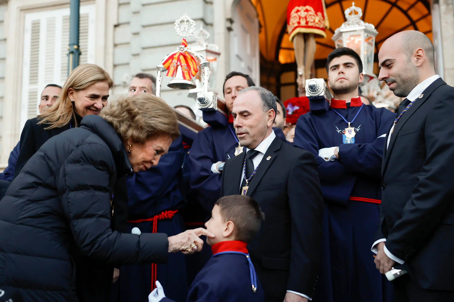La reina Sofía (i) saluda a Ismael, nieto del capataz Antonio Calahorro (c), en la salida de la procesión del Santísimo Cristo de los Alabarderos, este Viernes Santo en el Palacio Real, en Madrid