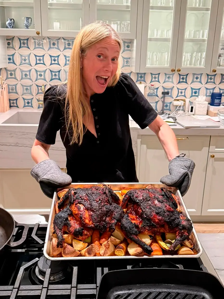 Gwyneth Paltrow en su cocina, mostrando una bandeja con dos pollos con patatas al horno.