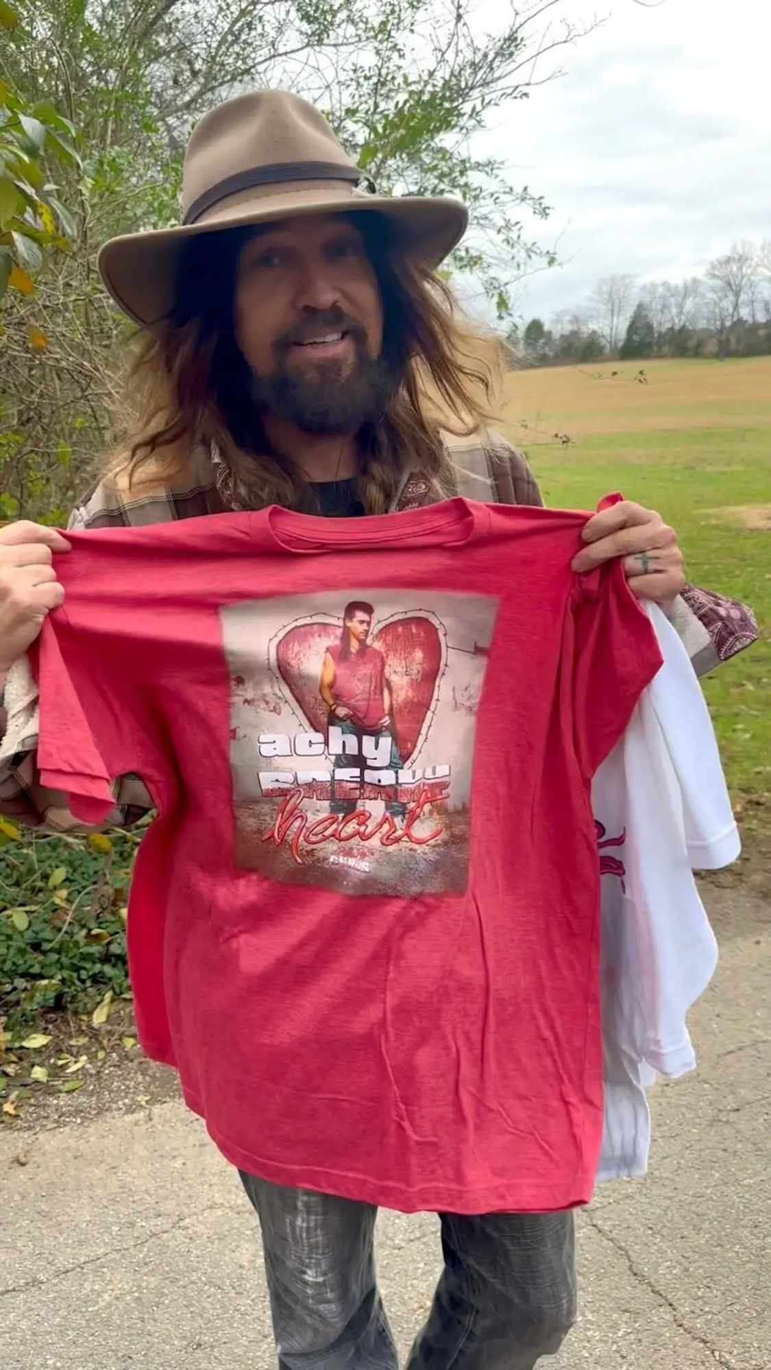 Billy Ray Cyrus sonriente, mostrando una camiseta.