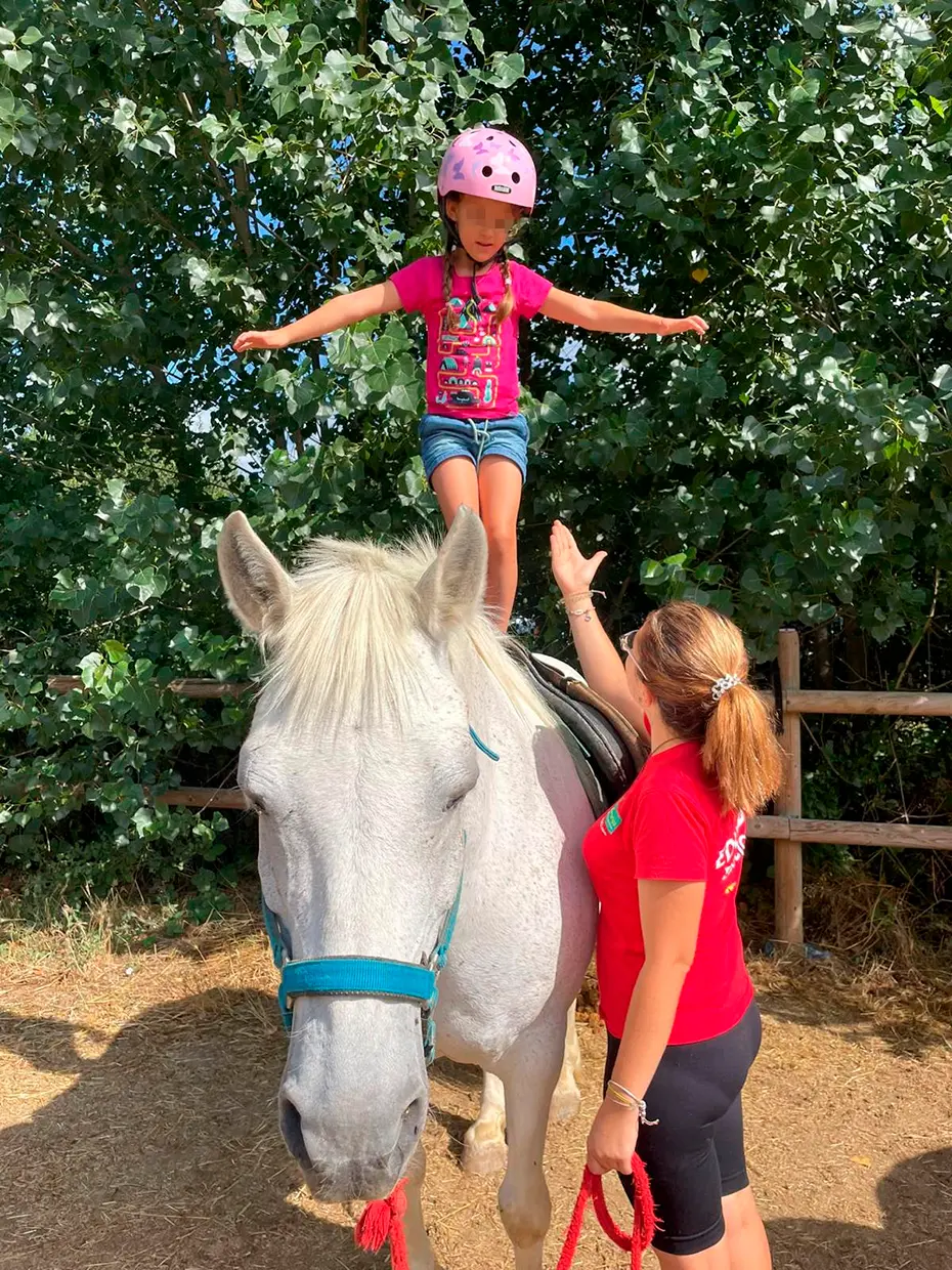 Niña de pie sobre un caballo, vigilada por una monitora